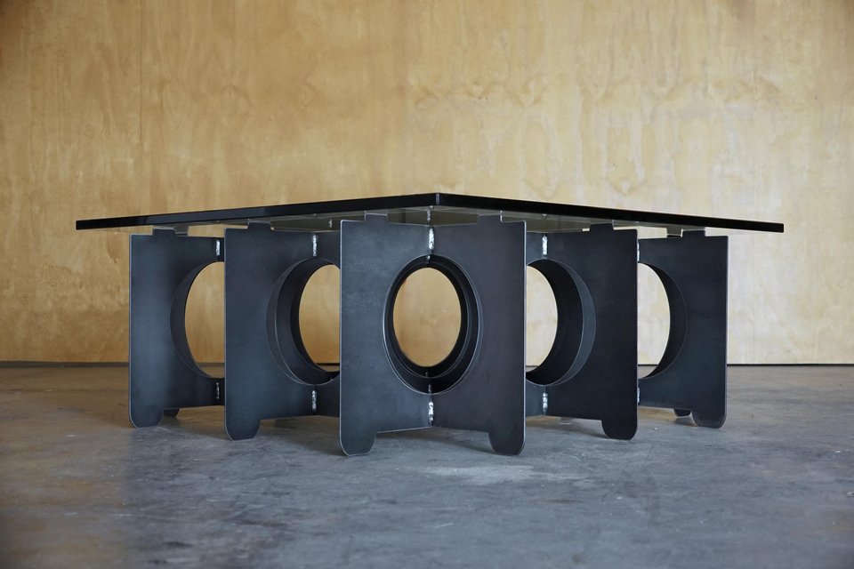 May include: A modern square coffee table with a black glass top. The base is constructed of dark gray metal panels with oval cutouts. The table's design is minimalist and industrial.