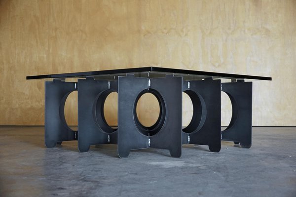 May include: A modern square coffee table with a black glass top. The base is constructed of dark gray metal panels with oval cutouts. The table's design is minimalist and industrial.